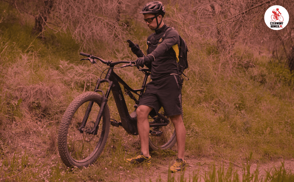 Rowerzysta w kasku i stroju sportowym korzysta ze smartfona przy czarnym rowerze elektrycznym w lesie – nawigacja GPS podczas jazdy e-bike’iem.